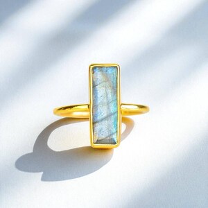 May include: A gold ring featuring a vertical rectangular labradorite gemstone. The stone displays iridescent blue and grey tones within a smooth gold setting. The band is thin and polished, creating a clean look that highlights the natural stone.