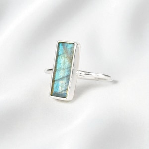 May include: A silver ring featuring a rectangular labradorite gemstone set in a smooth metal bezel. The stone displays a vibrant blue and gold iridescent flash across its surface, while the thin, polished silver band completes the design.
