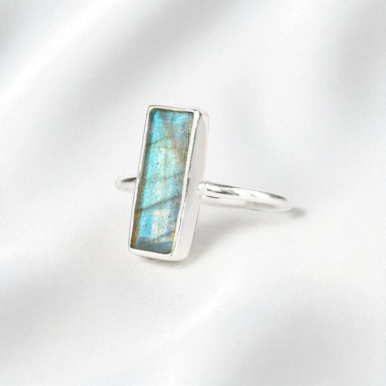 May include: A silver ring featuring a rectangular labradorite gemstone set in a smooth metal bezel. The stone displays a vibrant blue and gold iridescent flash across its surface, while the thin, polished silver band completes the design.