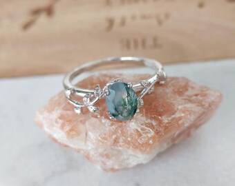 May include: A silver-toned ring featuring an oval-shaped moss agate gemstone with green inclusions. The band is designed with a delicate vine and leaf motif that wraps around the central stone, creating a nature-inspired look for this jewelry piece.