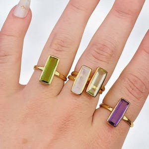 May include: Four gold-toned rings featuring rectangular, baguette-cut gemstones in various colors. The rings include a lime green stone, a pale pink stone, a light yellow stone, and a deep purple stone, each set in a simple, polished gold-colored band.