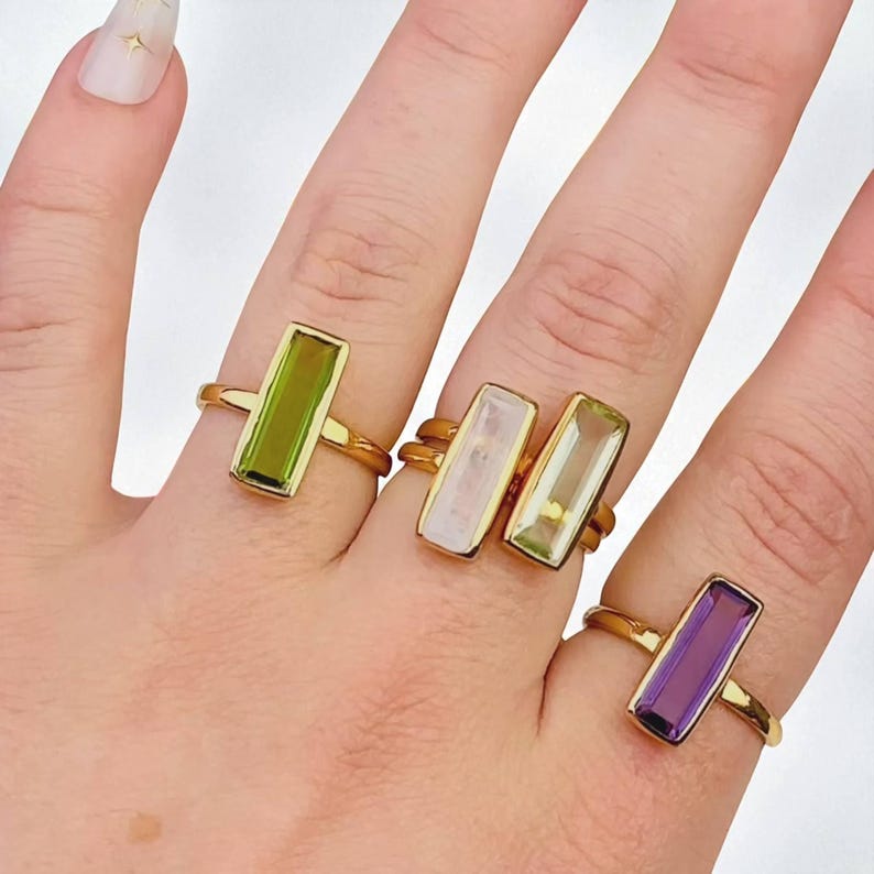 May include: Four gold-toned rings featuring rectangular, baguette-cut gemstones in various colors. The rings include a lime green stone, a pale pink stone, a light yellow stone, and a deep purple stone, each set in a simple, polished gold-colored band.