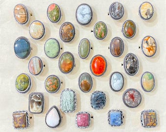 May include: A collection of twenty-nine rings featuring various polished gemstones in oval, rectangular, and teardrop shapes. Each stone is set in a decorative silver-toned metal frame with a twisted rope-like border detail around the edges.