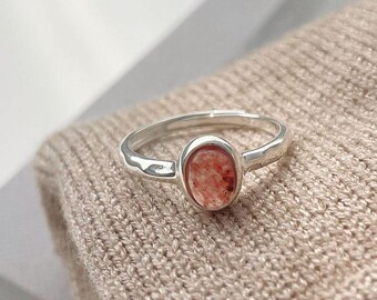 May include: A silver ring featuring a smooth, polished band and an oval-shaped, speckled peach and red stone setting. The ring has an open back design, visible at the top of the band, and is centered on a textured, light beige knit fabric surface.