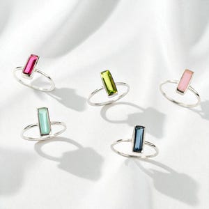 May include: Five silver rings, each featuring a single rectangular baguette-cut gemstone. The stones are colored in vibrant shades of hot pink, lime green, soft pastel pink, light mint green, and deep navy blue, all set on thin, polished silver bands.