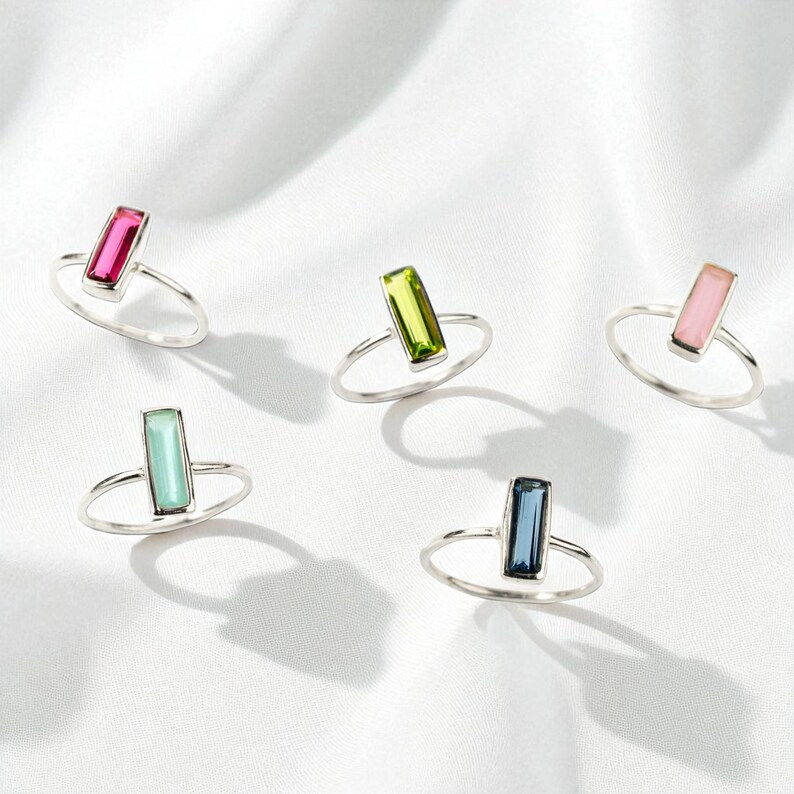 May include: Five silver rings, each featuring a single rectangular baguette-cut gemstone. The stones are colored in vibrant shades of hot pink, lime green, soft pastel pink, light mint green, and deep navy blue, all set on thin, polished silver bands.