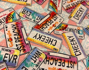 May include: Fabric with a repeating pattern of colorful license plates. The plates feature text like "JST PEACHY," "CHEEKY," and "SUNKISSD," with state names and rainbow designs. The background is a light pink and white geometric pattern.