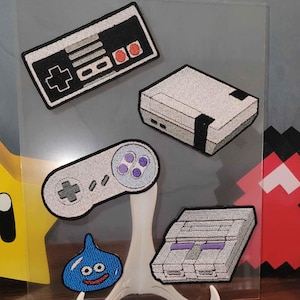 May include: A clear acrylic display featuring embroidered patches of retro video game controllers and consoles. Includes a controller, a console, and a blue slime character. A metal ruler measures approximately 12 inches.
