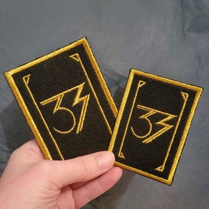 May include: Two black rectangular patches with gold embroidered borders and the number "33" in gold. The patches are held in a hand against a blue-grey background. The patches are likely for clothing or accessories.