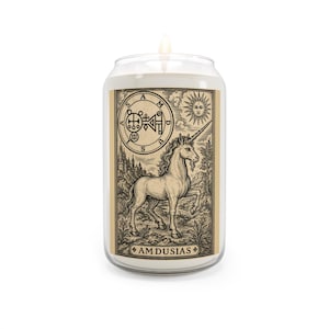 May include: A clear glass jar candle with a lit wick, featuring a beige rectangular label. The label displays a black ink illustration of a unicorn, a sun, and a circular sigil with the text AM DUSIAS printed at the bottom of the design.