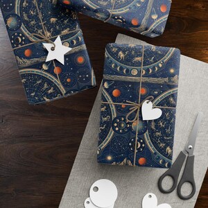 Könnte beinhalten: Zwei rechteckige Geschenke, eingewickelt in dunkelblaues Papier mit goldenen Himmelskarten, Sternbildern und orangefarbenen Planeten. Jedes Paket ist mit brauner Schnur verschnürt und mit einem weißen Papieranhänger versehen.