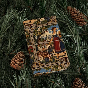 May include: A rectangular gift wrapped in paper featuring a Bavarian-themed illustration of a person in traditional clothing holding a beer stein, surrounded by pretzels and edelweiss flowers, tied with thin twine and placed among pine needles.