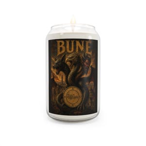 May include: A clear glass jar candle with a lit wick, featuring a label with the text BUNE in gold lettering. The label displays a dark, detailed illustration of a three-headed creature with wings and a circular sigil symbol in the centre.