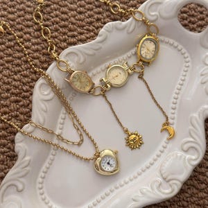 May include: A gold-toned necklace featuring three integrated watch faces connected by chain links, with dangling sun and moon charms. A separate gold-toned necklace features a heart-shaped watch pendant on a delicate beaded chain.