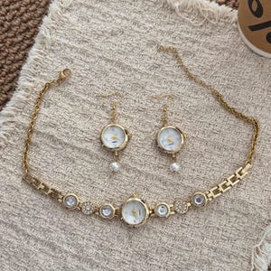May include: A gold-toned jewelry set consisting of a necklace and two matching earrings. The necklace features a central watch face with a white dial, flanked by circular links with clear crystals. The earrings are miniature watch faces with white dials and dangling white pearls.