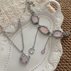 Può includere: Un set composto da collana e bracciale color argento con diversi quadranti di orologio in rosa pallido. La collana presenta un ciondolo a forma di cuore su una catenina a palline, mentre il bracciale unisce tre quadranti ovali con vari ciondoli.