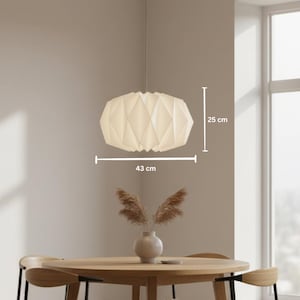 May include: A white pendant light with a geometric, folded paper-like structure. The lamp has a height of 25 cm and a width of 43 cm, as indicated by the white measurement lines and text visible on the image, suspended from a thin cord.