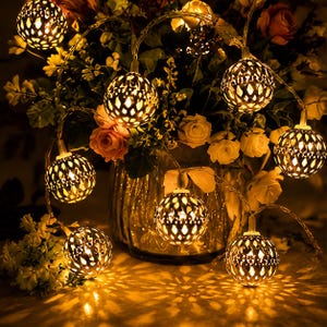 May include: A string of decorative lights featuring spherical metal globes with intricate cut-out patterns. Each globe emits a warm, golden glow, casting detailed shadows. The lights are arranged in a cluster, illuminating the surrounding area.