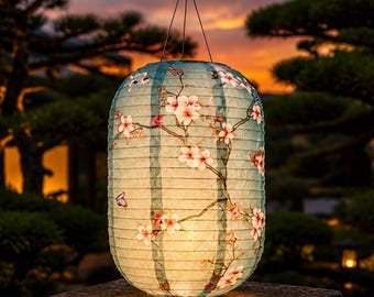May include: A cylindrical paper lantern with a light blue background featuring a printed pattern of pink cherry blossoms and small butterflies. The lantern has a ribbed texture and is suspended by a thin metal wire hook at the top.