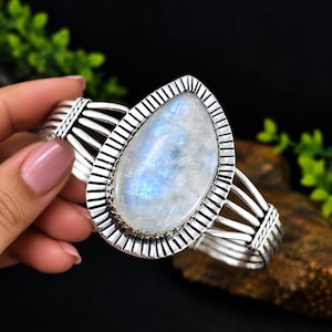 Op de afbeelding: Een zilveren manchetarmband met een grote, peervormige maansteen cabochon in het midden. De steen heeft een doorschijnend wit uiterlijk met blauwe iridiscentie. De metalen band heeft een geribbeld frame en meerdere zijdelingse draden.