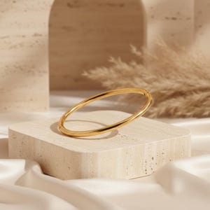 May include: A polished gold-tone bangle bracelet with a smooth, rounded surface and a circular shape. The bracelet features a simple, continuous design with a reflective metallic finish that catches the light, creating a clean and uniform appearance.