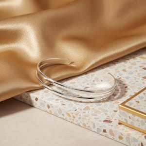 May include: A polished silver cuff bracelet featuring a double-band design with a smooth, reflective finish. The open-ended bangle is constructed from two parallel, thin silver wires that curve to form a circular shape for the wrist.