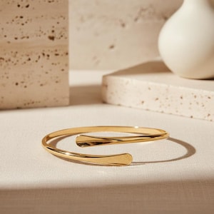 May include: A gold-toned open cuff bracelet with a polished, reflective finish. The bracelet features a bypass design with two slightly flattened, rounded ends that overlap slightly, creating a sleek and continuous circular silhouette.