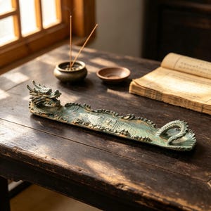 May include: A long, narrow incense burner shaped like a dragon with a weathered, verdigris-coloured finish. The dragon head is at one end, and the tail curls at the other, forming a trough for incense sticks. The surface features detailed scales.