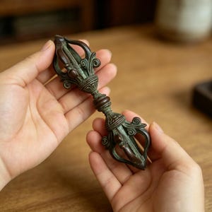 May include: A bronze-coloured metal vajra ritual object featuring a central grip with ribbed detailing and two symmetrical ends composed of four curved prongs that meet at a pointed tip, showcasing an aged, dark patina with intricate scrollwork.