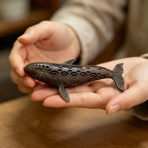 Könnte beinhalten: Eine dunkelbraune Walfigur aus Metall mit einem komplizierten durchbrochenen Design aus sich wiederholenden Wellenmustern am Körper. Die Skulptur hat einen glatten, abgerundeten Kopf mit kleinen kreisförmigen Perforationen und eine Schwanzflosse.