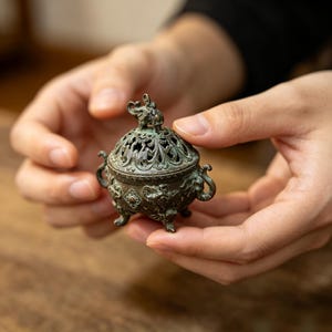 Puede incluir: Un pequeño quemador de incienso de metal en tono bronce oscuro con una tapa de filigrana intrincada rematada por una figura de elefante en miniatura. El recipiente redondo tiene tres patas, asas ornamentadas y motivos de elefantes en relieve.