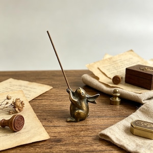 May include: A small, antique-style brass figurine of a rabbit standing on its hind legs with its front paws raised to hold a single, thin, brown incense stick. The rabbit has a smooth, metallic finish with dark, aged patina accents.