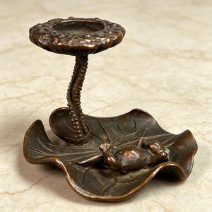 Puede incluir: Un portavelas de metal en tono bronce con una base en forma de hoja de nenúfar grande sobre la que descansa una pequeña figura de rana. Un soporte texturizado en forma de tallo se eleva desde la base para sostener una plataforma circular con bordes festoneados.