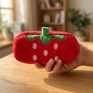 Peut inclure: Une trousse à crayons rectangulaire en peluche en forme de fraise, dotée d'un corps en tissu rouge vif avec des graines blanches brodées et une tige en tissu vert sur le dessus. La trousse comprend une petite chaînette en métal sur la fermeture.