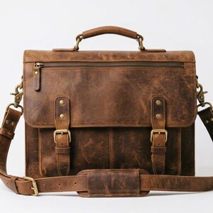 May include: A brown leather briefcase featuring a top handle, a detachable shoulder strap with a padded section, and a front flap secured by two buckle straps. The bag includes a horizontal zippered pocket on the front flap and brass hardware.