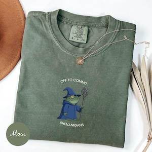 May include: A moss green t-shirt featuring a graphic of a frog wearing a blue wizard hat and robe while holding a staff. The text above the frog reads "OFF TO COMMIT" and the text below reads "SHENANIGANS". The shirt has a Comfort Colors label.