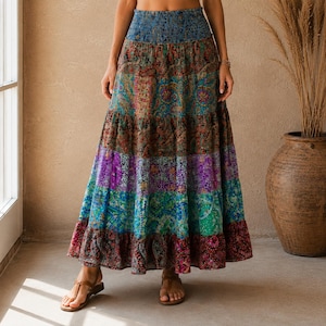 Puede incluir: Una falda larga de capas con cintura elástica fruncida y un estampado de cachemira multicolor vibrante. La falda muestra bandas horizontales con patrones variados en tonos azul, morado, verde, marrón y rojo, llegando hasta los tobillos.