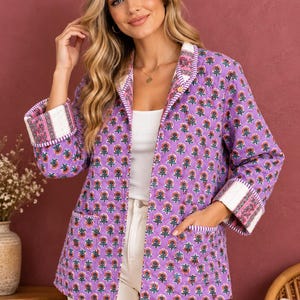 May include: A quilted jacket in a vibrant purple hue featuring a repeating floral pattern with orange and green details. The jacket has long sleeves with folded cuffs, a front opening, and two patch pockets, all finished with striped trim.