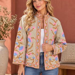 May include: A beige open-front jacket featuring a mandarin collar and long sleeves. The garment is decorated with embroidered floral patterns in vibrant pink and light blue, accented by brown vine-like stems along the front and sleeves.