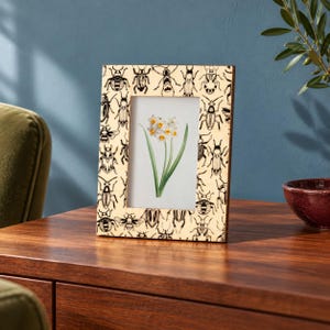 May include: A rectangular picture frame with a light beige border featuring a repeating black ink illustration pattern of various insects. The center of the frame displays a botanical print of white and yellow daffodil flowers with green stems.