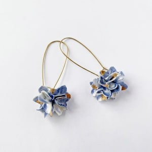 Puede incluir: Un par de pendientes colgantes con racimos de cuentas de cerámica en forma de flor azul y blanca. Cada racimo está acentuado con pequeños toques de pintura dorada y cuelga de un gancho de alambre de metal dorado fino y curvado.