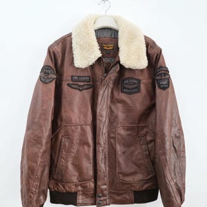 Peut inclure: Un blouson aviateur en cuir marron doté d'un col en fausse fourrure crème, de poignets et d'un ourlet côtelés marron foncé, ainsi que de plusieurs écussons brodés avec le texte PME Legend et American Classic sur la poitrine et les manches.