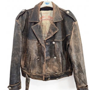 May include: A distressed brown leather jacket featuring a double-breasted front with silver-toned metal buttons, shoulder epaulets, and a belted waist. The interior lining displays a printed map graphic with the text "WORLD TRAVELER".