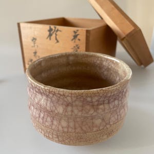 May include: A ceramic tea bowl with a light beige base and a prominent purple crackle glaze finish. Behind the bowl sits a square wooden storage box with a separate lid, featuring black ink calligraphy characters on the front side of the box.