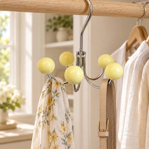 May include: A chrome-plated metal hook with four arms, each ending in a smooth, pale yellow spherical knob. The hook features a curved top for hanging on a rod and is currently holding a floral print scarf and a beige leather strap accessory.