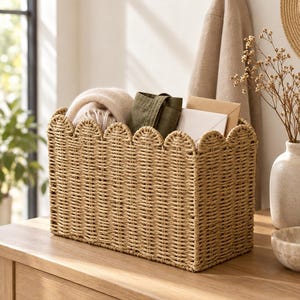 May include: A rectangular woven storage basket made of light tan natural fibers, featuring a decorative scalloped top edge. The basket is filled with a folded beige textile, a dark olive green fabric item, and several white and tan paper envelopes.