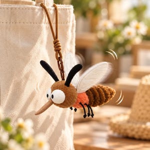 Op de afbeelding: Een pluche muggen-sleutelhanger met een bruine kop en lichaam, een feloranje middengedeelte, grote witte ogen, zwarte voelsprieten en doorschijnende witte vleugels. Het insect hangt aan een bruin koord aan een beige canvas tas.