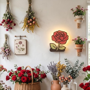 May include: A wall-mounted rose-shaped light fixture with a red flower and green leaves, glowing with a warm backlight. The design features bold black outlines, a central red rose head, and a dark base, connected to a white power cord.