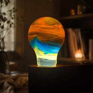 May include: A light bulb shaped decorative object with swirling orange, blue, and yellow colors inside the glass. The text "Solar system" is printed above the bulb, which is set into a dark, smooth, rounded wooden base on a flat surface.
