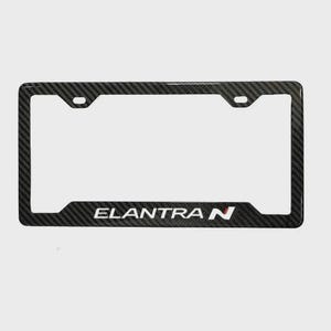 May include: A rectangular license plate frame featuring a black and gray carbon fiber pattern. The bottom edge displays the white text ELANTRA followed by a red and white N logo. The frame includes two mounting holes at the top for attachment.